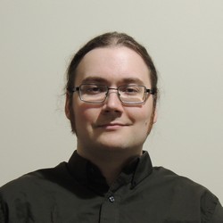 Chris smiling; he
			has curly hair, mutton chops, glasses, and is wearing a solid brown
			button-up shirt